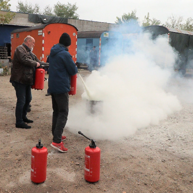 Zwei Mitarbeitende der gemeinnützigen C.U.B.A. GmbH sind gerade dabei, mithilfe eines Handfeuerlöschers, ein Feuer in einer 200 l Blechtonne zu löschen.