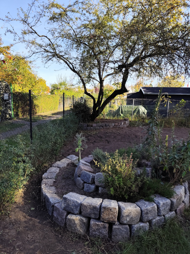 Im Vordergrund befindet sich eine Pflanzschnecke aus behauenen Natursteinen. Im Hintergund rahmt eine zweilagige Trockenmauer – ebenfalls aus behauenen Natursteinen – einen Baum ein.